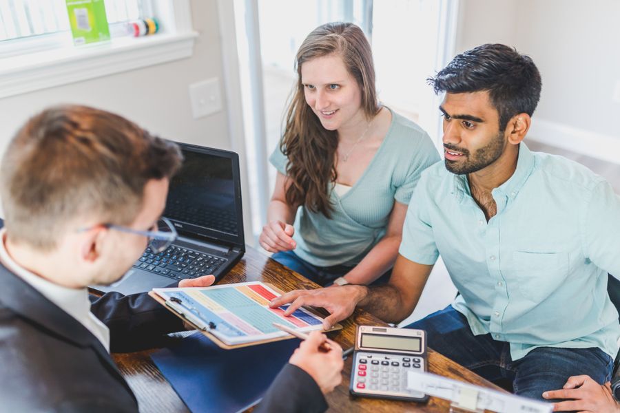 A couple sitting with a broker at a table covered in paperwork and a calculator, discussing buying a home