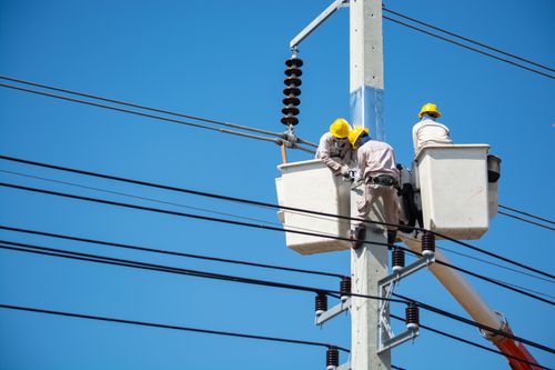 Two linesmen working from a bucket lift on a power pole against a blue sky